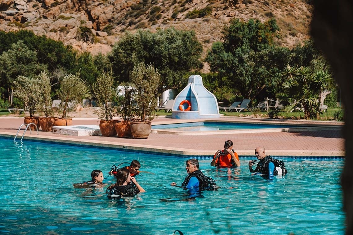 Mensen in duikuitrusting oefenen in een zwembad met op de achtergrond een heuvel, bomen en een kleiner zwembad.