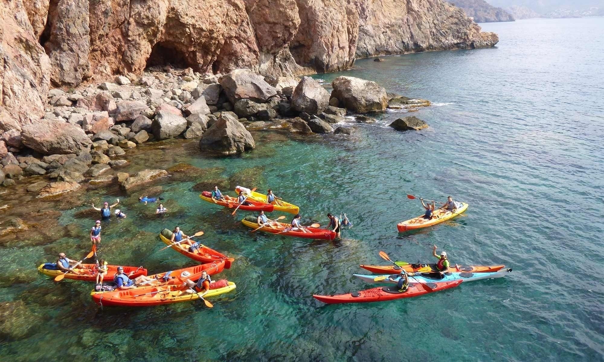 Varias personas disfrutan del kayak y la natación en aguas turquesas y claras junto a imponentes acantilados rocosos.