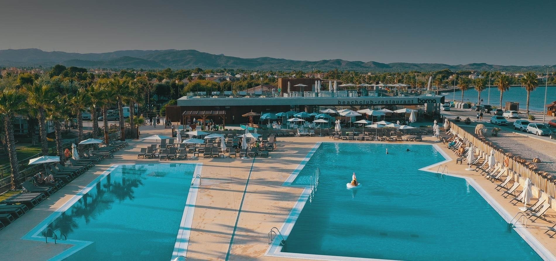 a large swimming pool surrounded by chairs and umbrellas in front of a beachclub restaurant