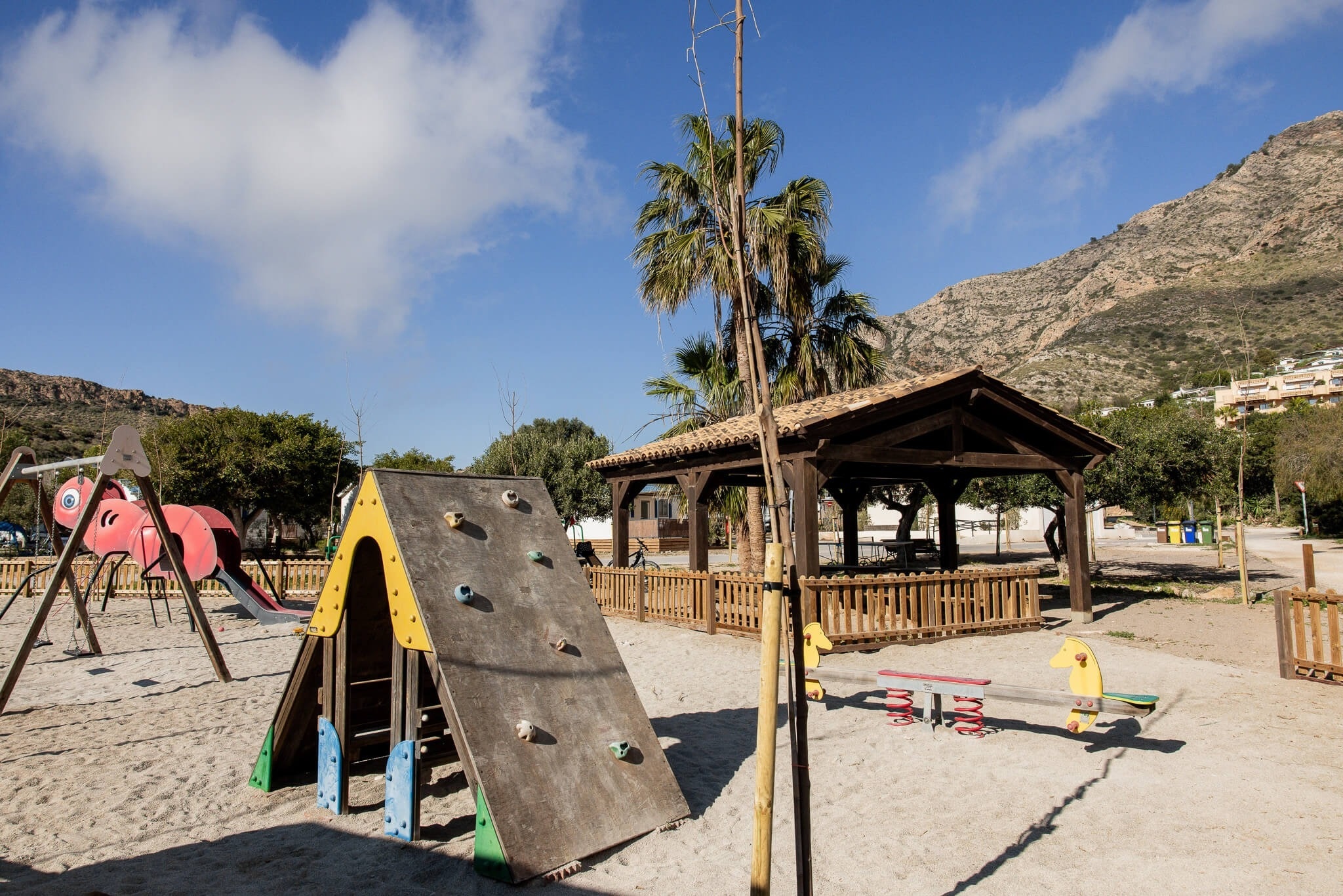 Un parque infantil de arena con varias estructuras de juego, una pérgola de madera y montañas al fondo bajo un cielo azul con nubes.