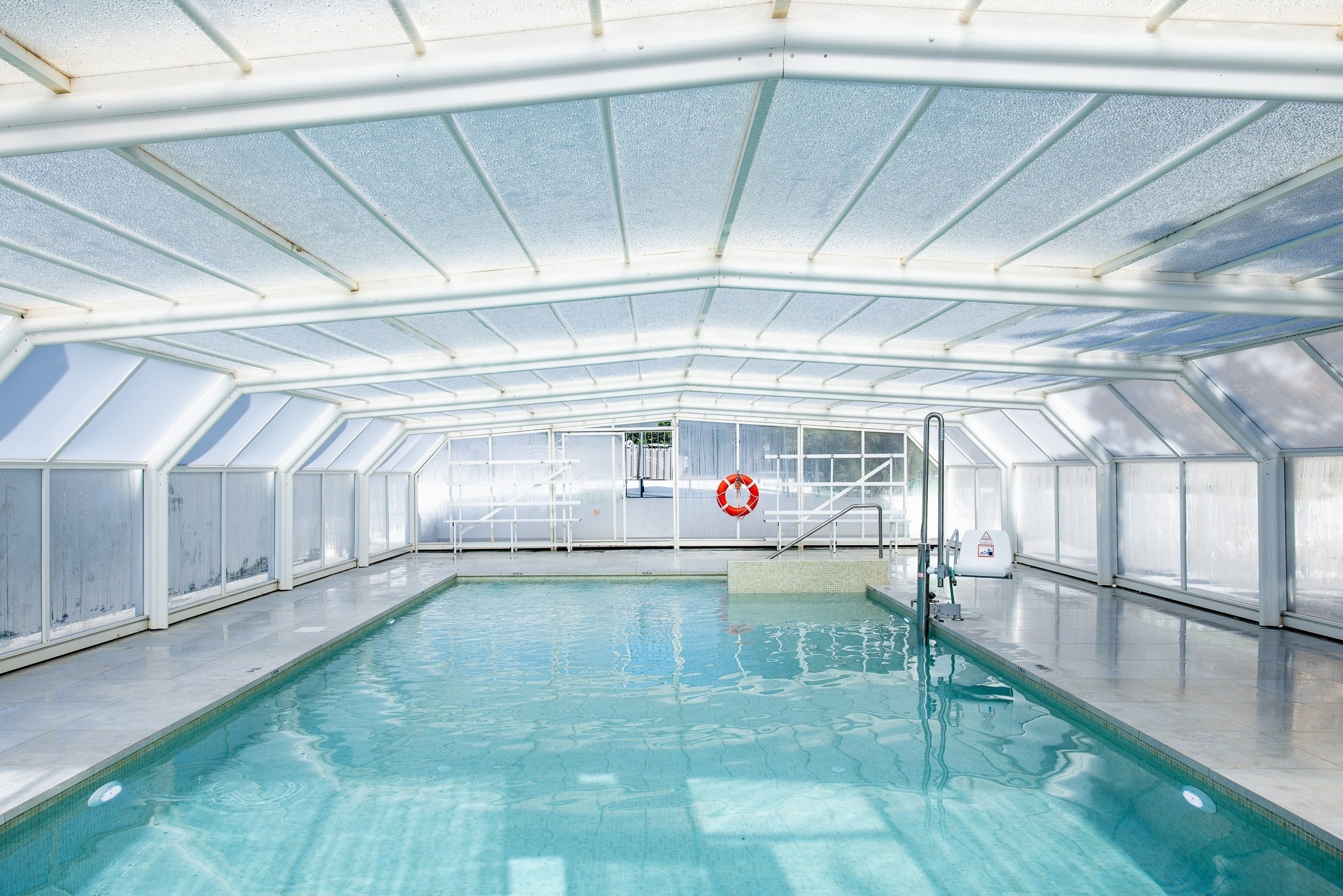 Una piscina cubierta y luminosa con un cerramiento translúcido y un salvavidas naranja visible al fondo.