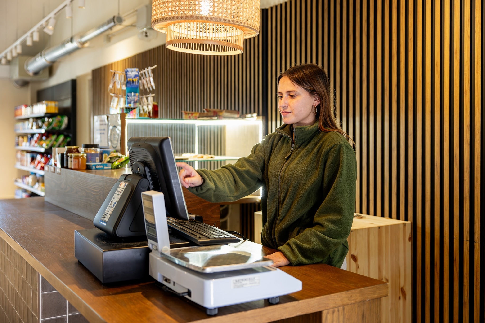 Een jonge vrouw in een groene fleece trui bedient een kassasysteem met een weegschaal op een houten toonbank in een winkel.