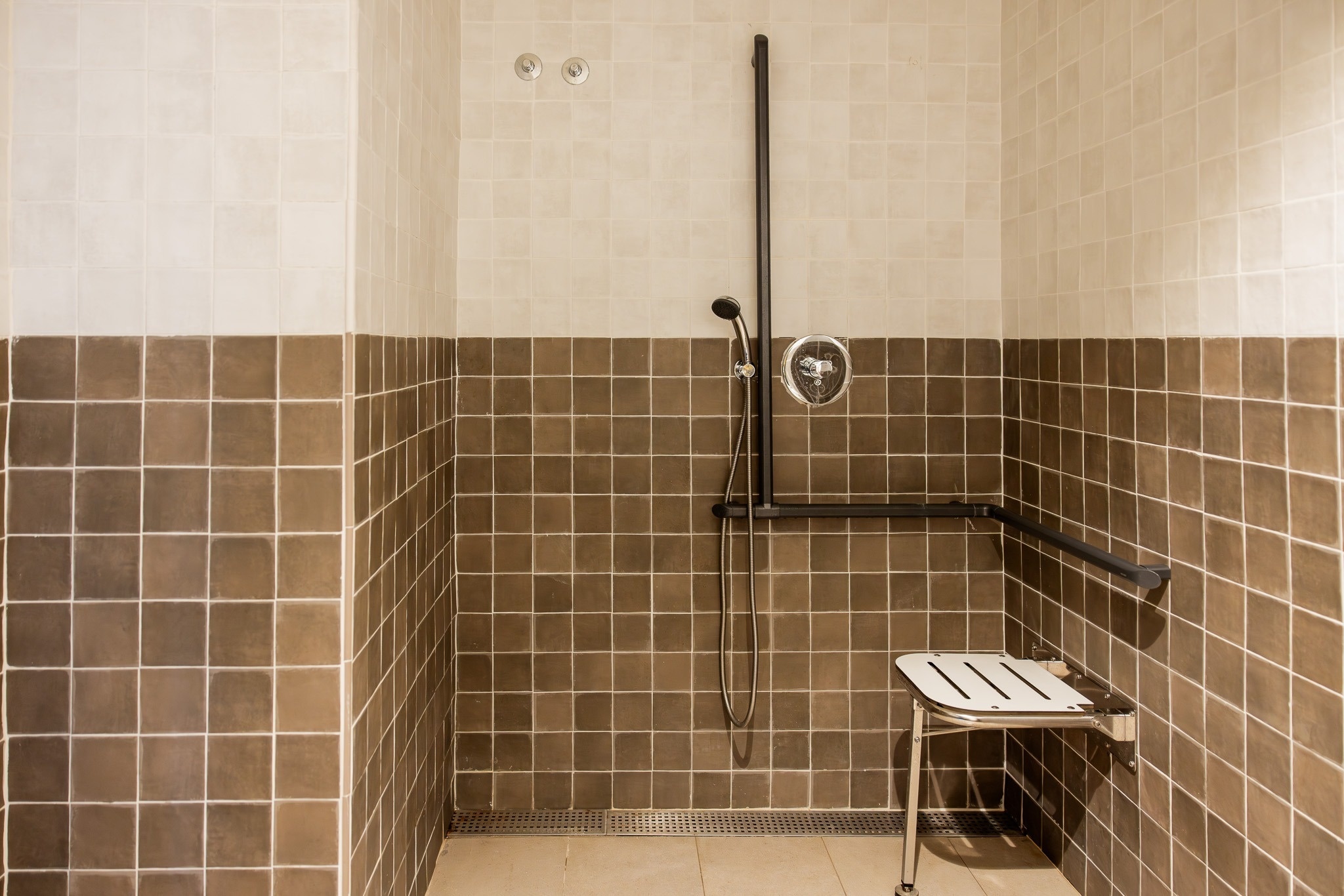 Een toegankelijke doucheruimte met crème en bruine tegelwanden, uitgerust met een douchekop, handgrepen en een opklapbare zitting.