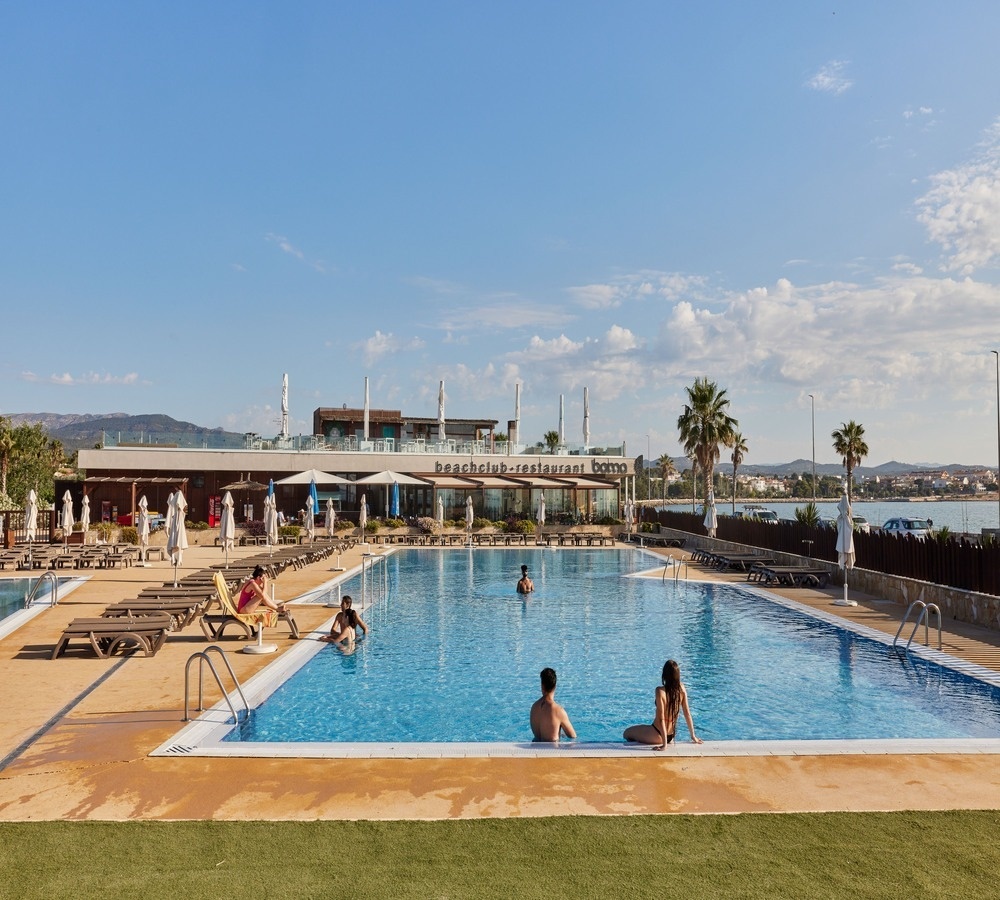 people are swimming in a large pool in front of a beach club restaurant