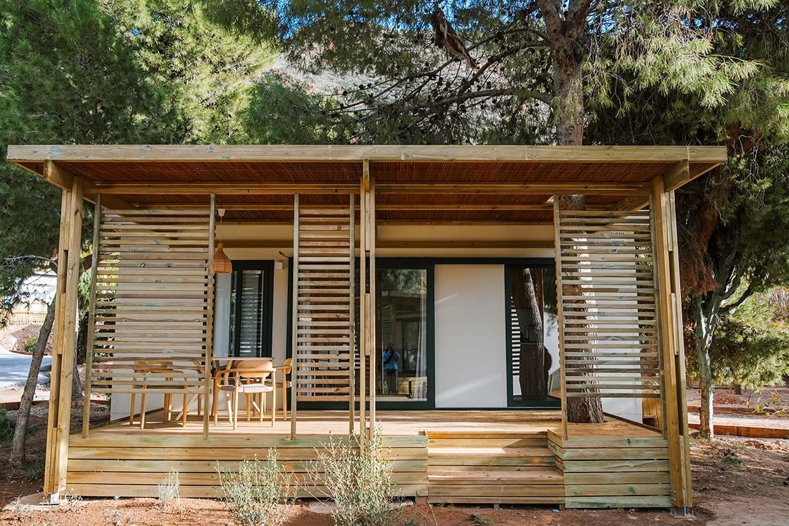 Een moderne houten bungalow met een veranda van houten lamellen en een overkapping van geweven matten, omringd door dennenbomen.