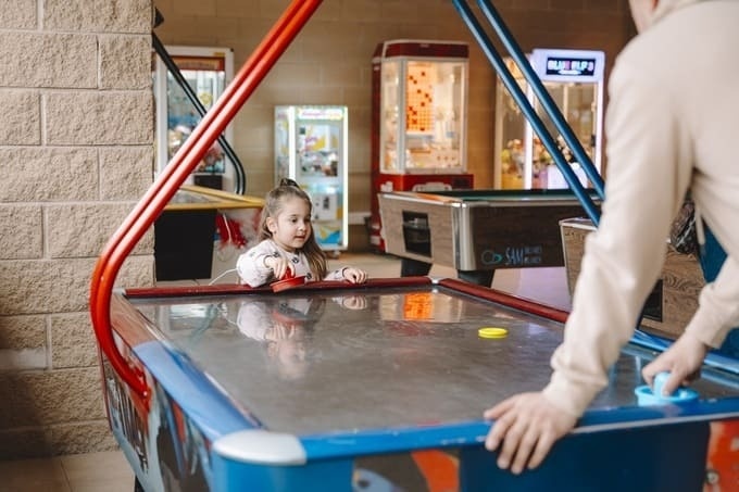 Niña y adulto jugando air hockey en sala de juegos de hotel.