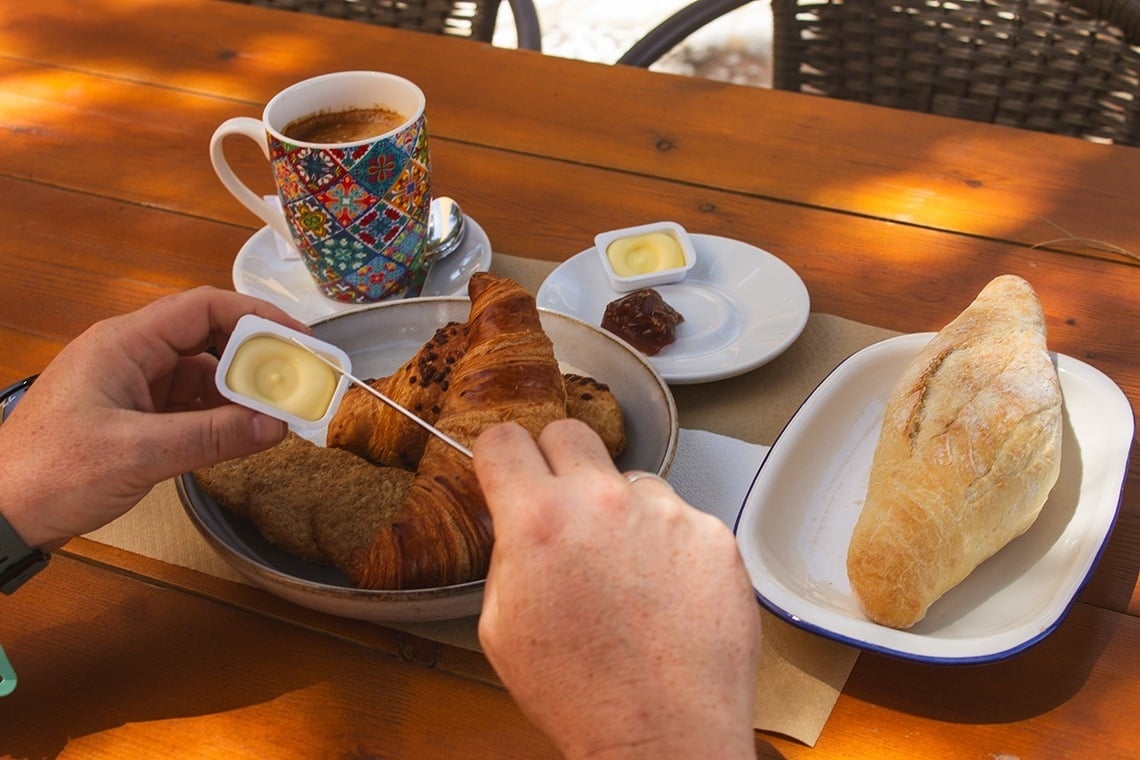 a person spreading butter on a croissant next to a cup of coffee