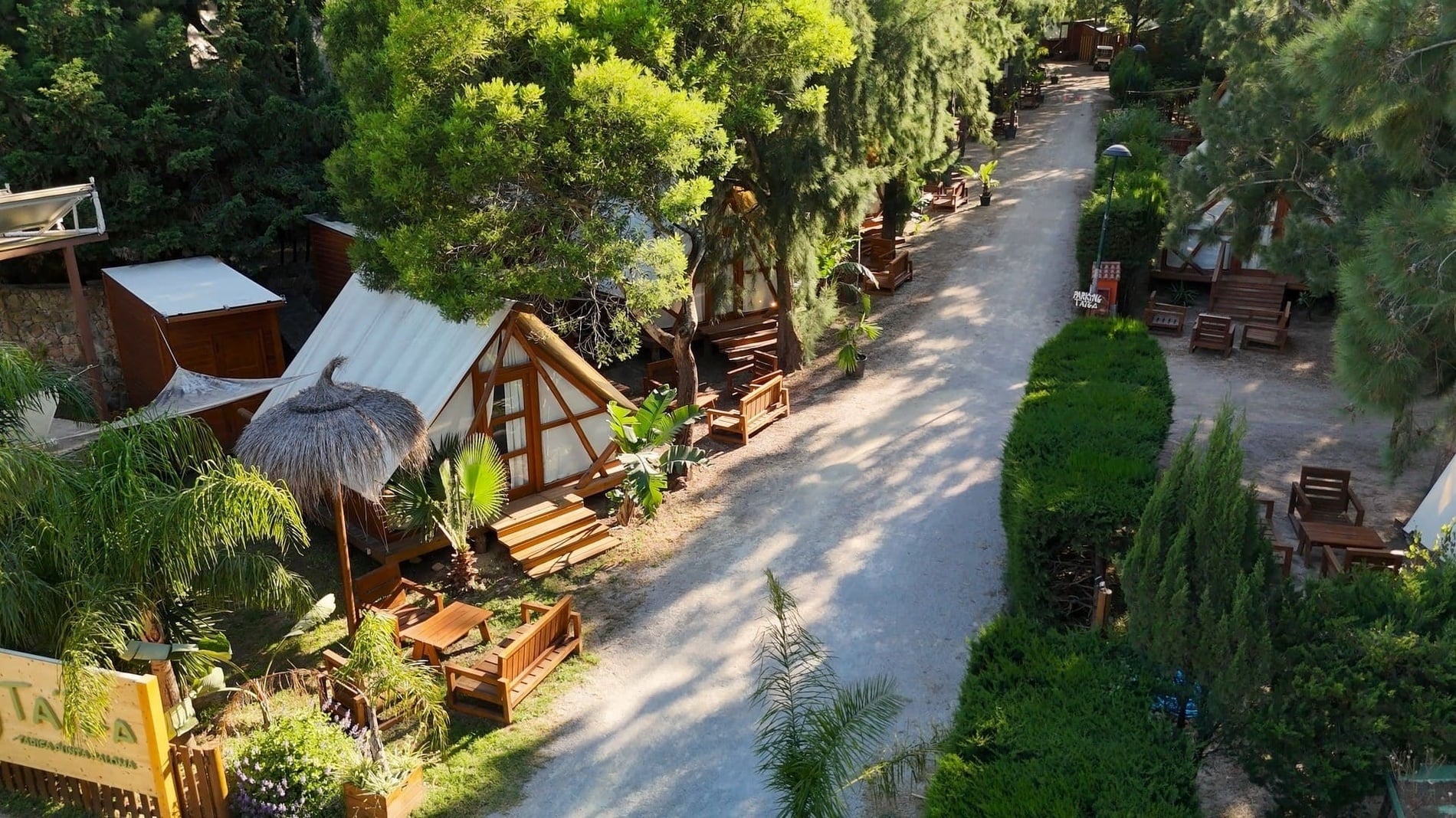 Un camino de tierra serpentea a través de un complejo de cabañas de madera y tiendas de campaña tipo glamping, rodeado de densa vegetación y árboles.