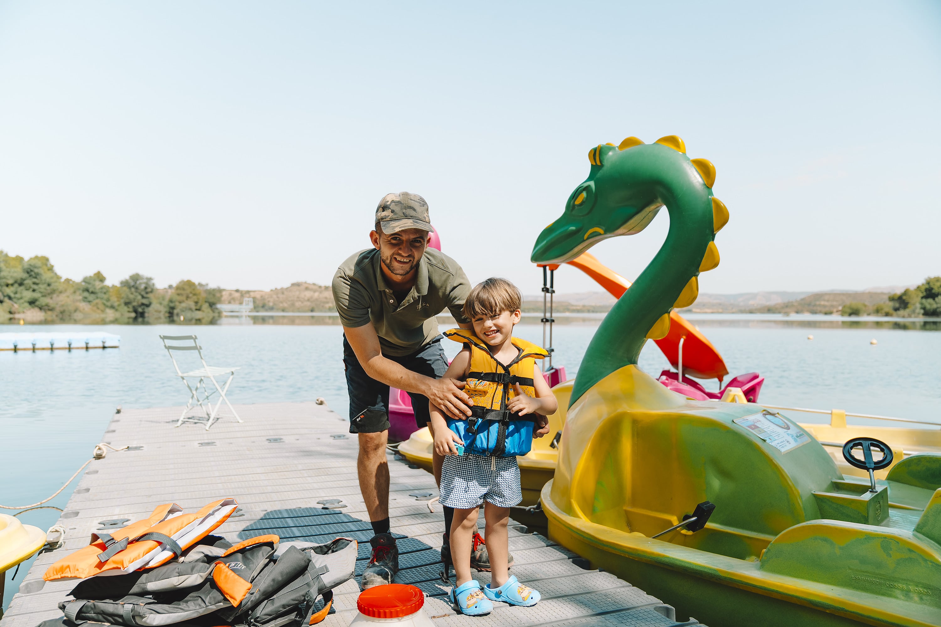 Padre e hijo con chalecos salvavidas junto a un bote de pedales con forma de dragón en un hotel TAIGA Lake Caspe | TAIGA | Campings & Resorts en España