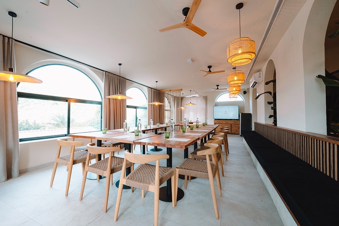 Een moderne vergaderruimte met een lange houten tafel, stoelen met rieten zittingen, grote boogvensters en houten plafondventilatoren en hanglampen.