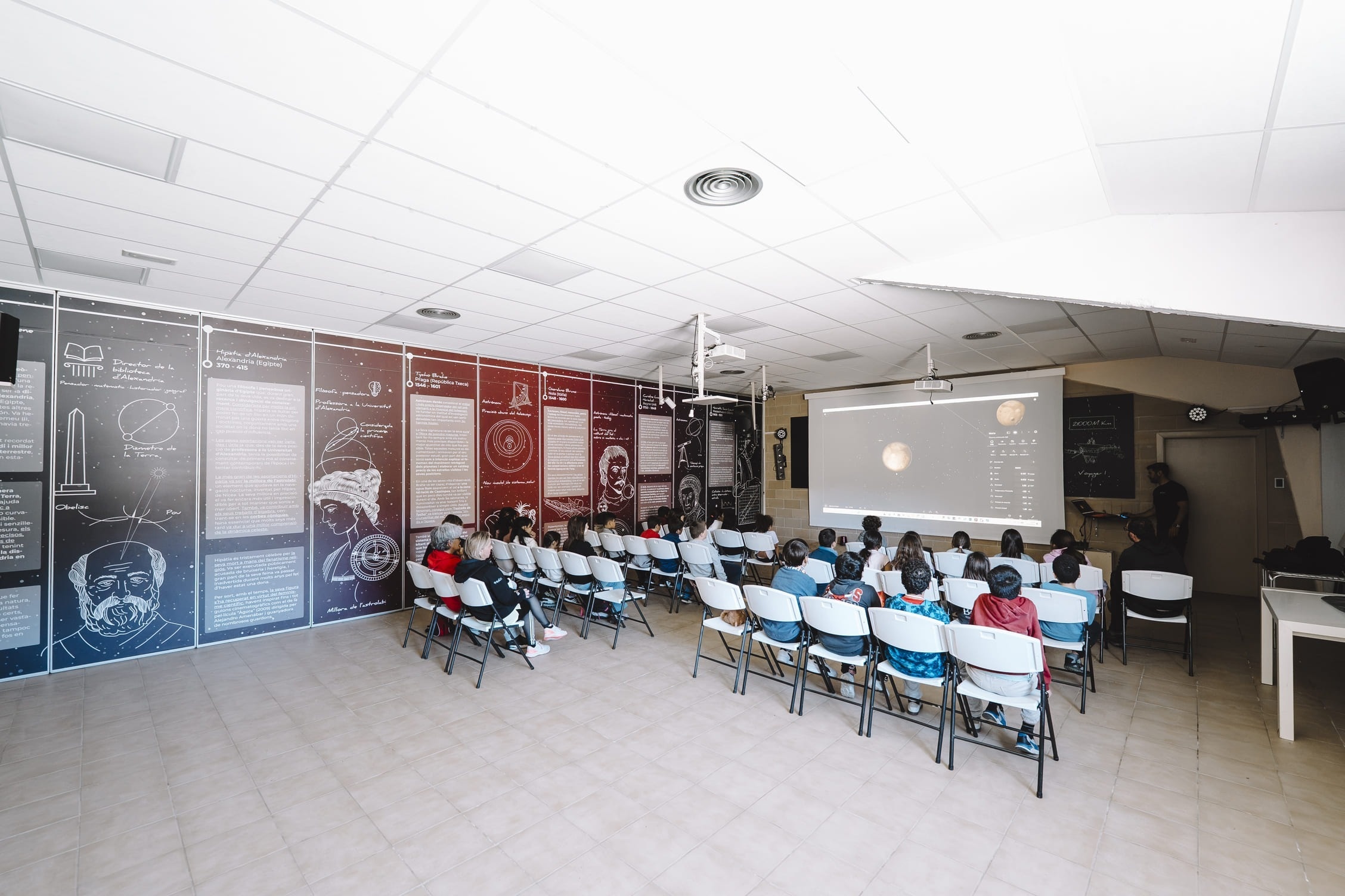 Niños aprendiendo ciencia en aula con proyección de planetas y murales históricos. Actividad educativa familiar.