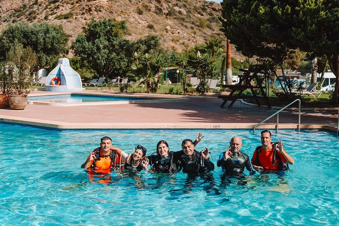Een groep van zes duikers, gekleed in wetsuits en met maskers, poseert lachend in een zwembad en maakt overwegend het 