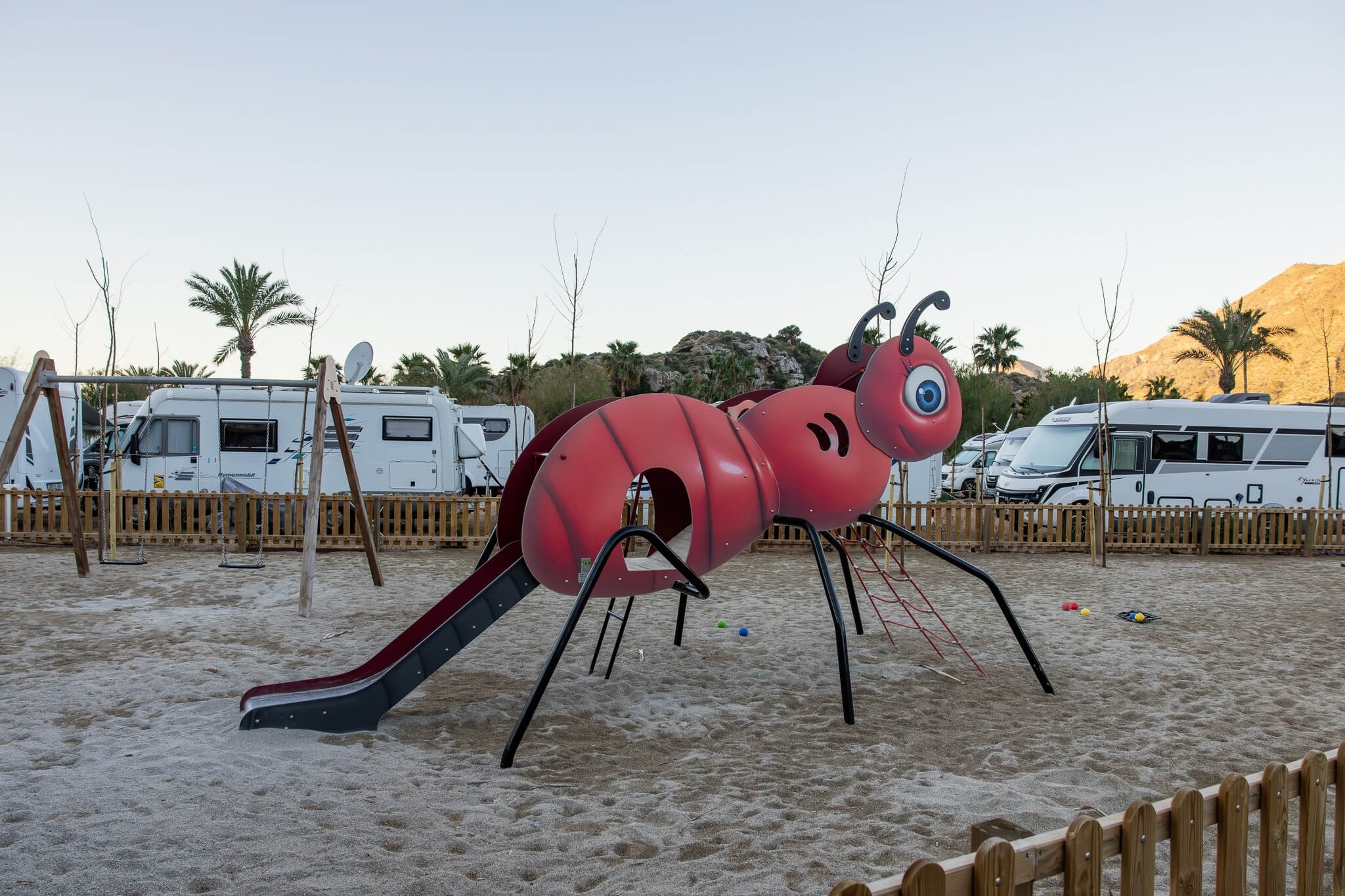 Un parque infantil con un tobogán rojo en forma de hormiga gigante se encuentra en un arenero, rodeado de autocaravanas, palmeras y montañas al fondo.