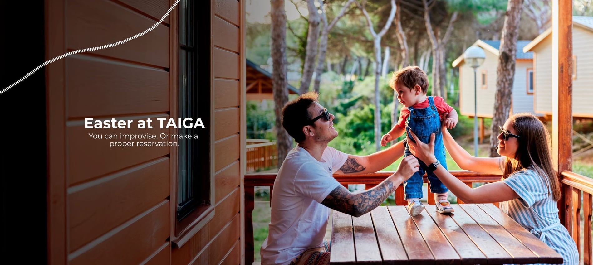 A family with a young child enjoys time together on the porch of a wooden cabin, surrounded by trees and other cabins in the background.
