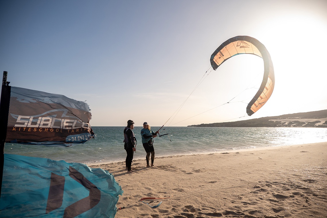 Kitesurf Taiga: Corporativa en Tarifa, instructor con cometa naranja y alumnos en la playa.