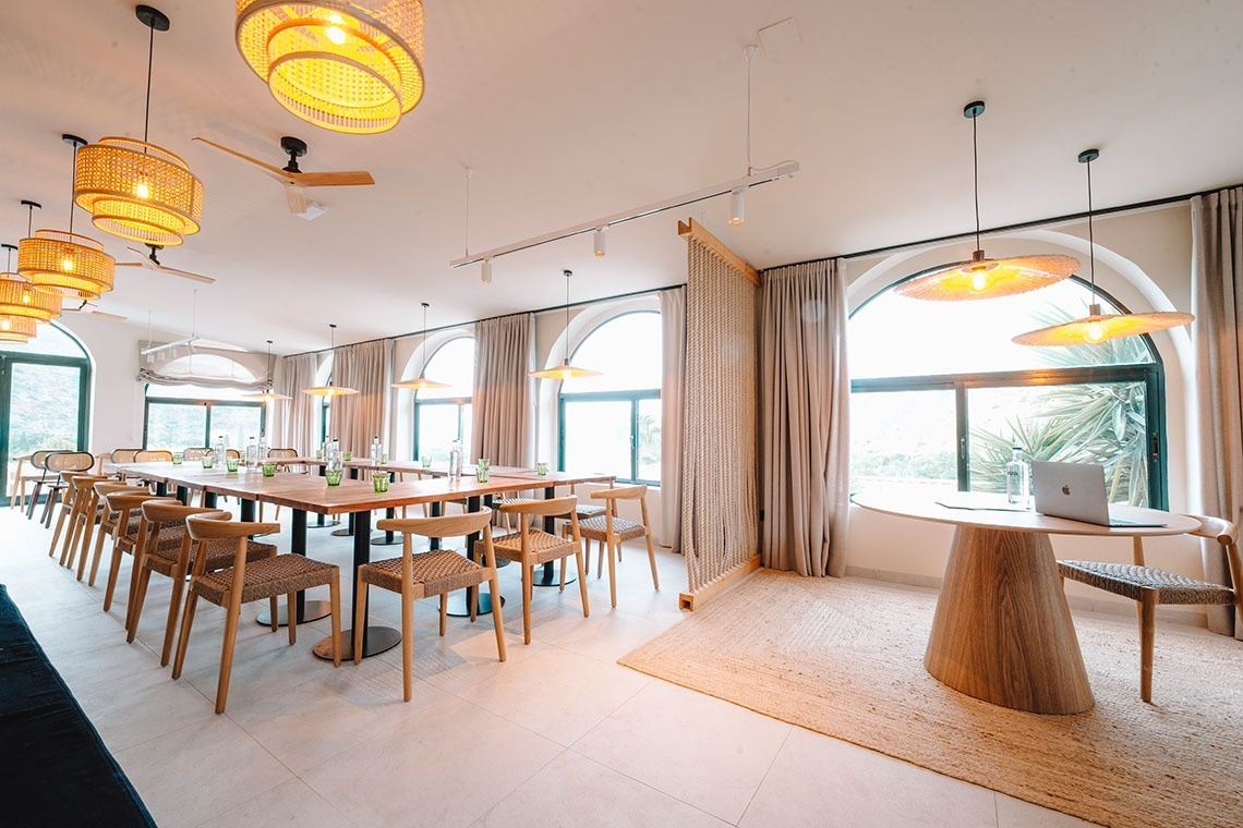 Een lichte en moderne vergader- of evenementenruimte met houten tafels en stoelen, rieten hanglampen, grote boogvensters en een afgescheiden werkplek met een laptop.