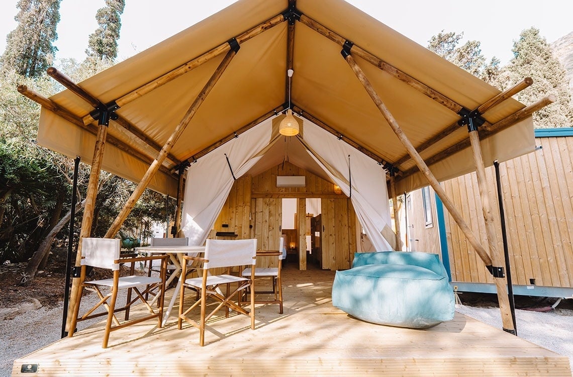 Een glampingtent met een houten terras, comfortabel meubilair en een inkijkje in het houten interieur staat in een groene omgeving.