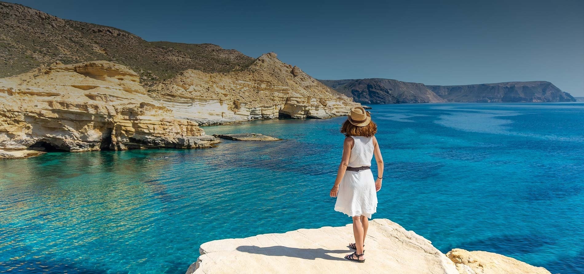 una mujer con un vestido blanco y un sombrero está de pie en un acantilado mirando al océano