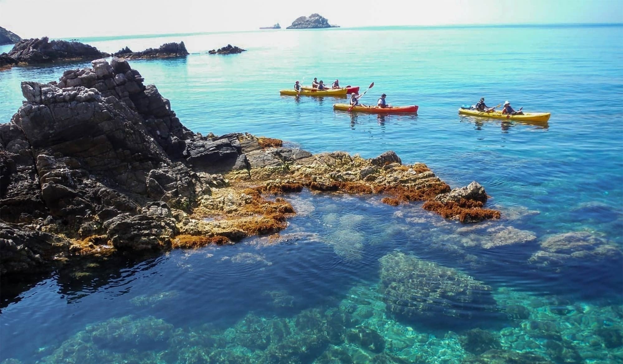 Varias personas disfrutan del kayak y la natación en aguas turquesas y claras junto a imponentes acantilados rocosos.