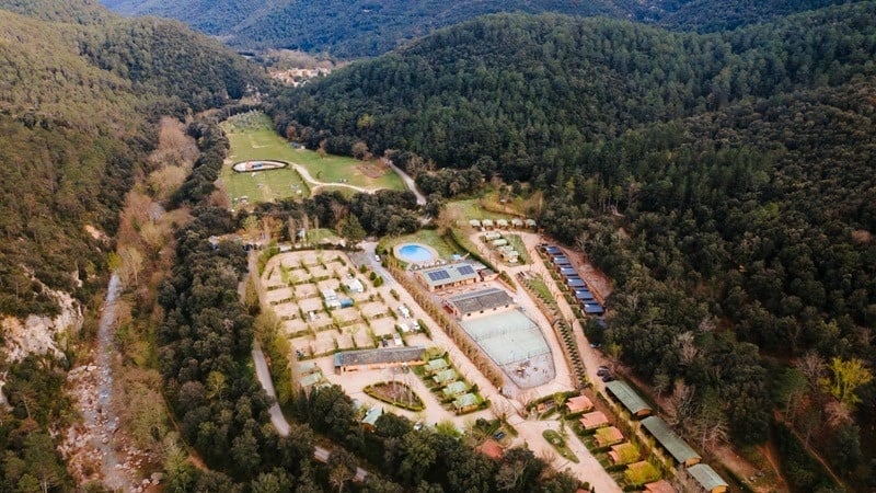 Complejo turístico en valle de montaña con río, piscina, bungalows y parcelas de camping.