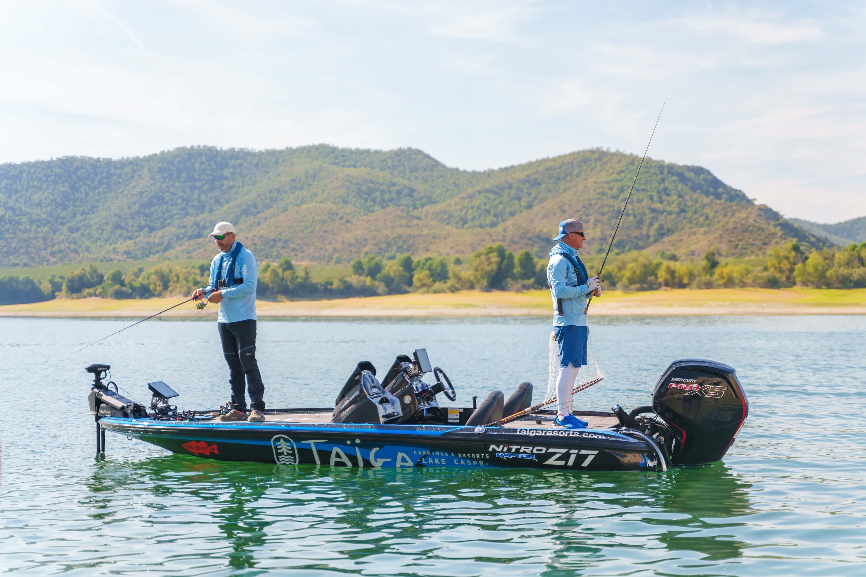 Dos pescadores disfrutan de un día de pesca desde una lancha especializada en un lago sereno, con montañas verdes de fondo.