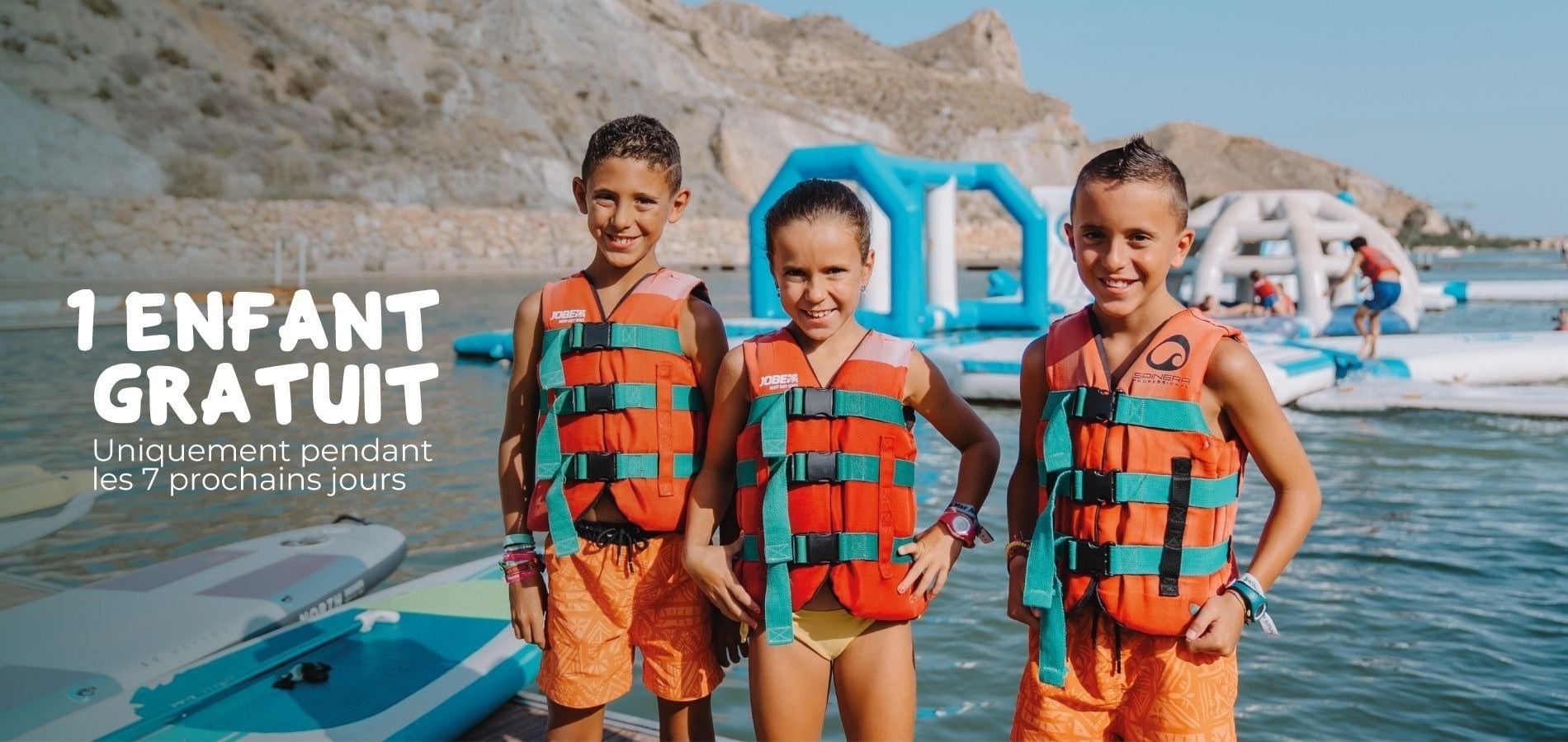 Trois enfants souriants en gilets de sauvetage devant un parc aquatique. Offre 1 enfant gratuit.