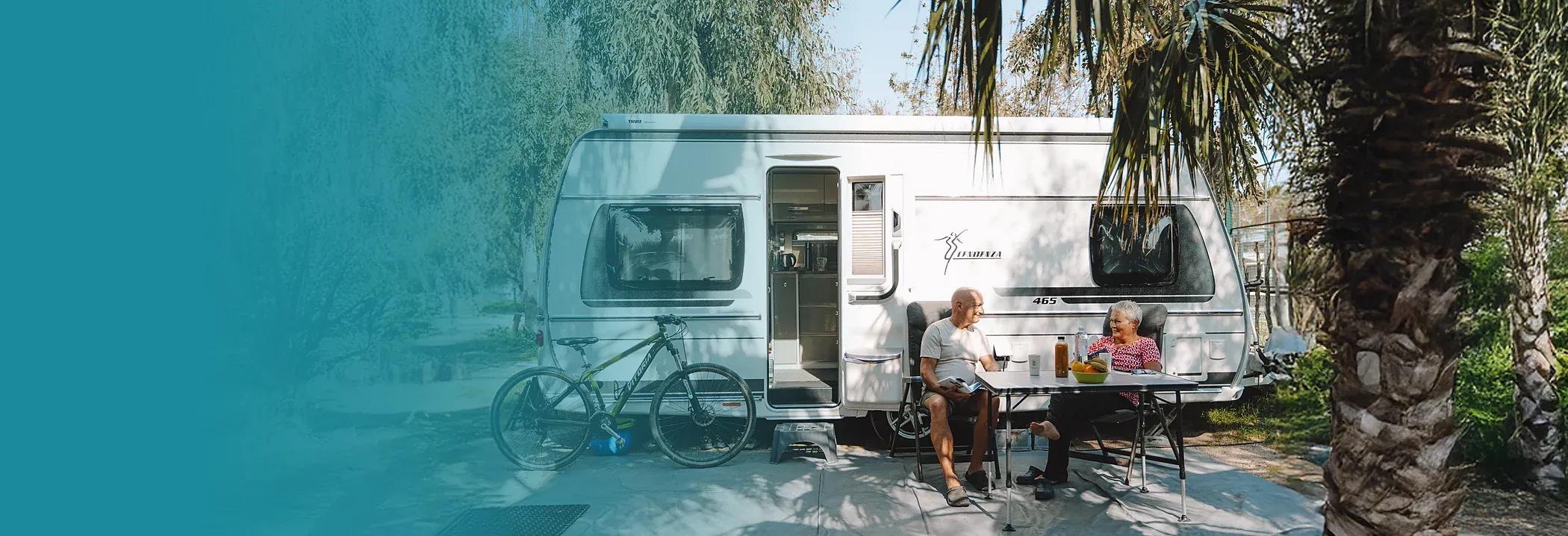 Pareja sentada a la mesa junto a su caravana en una parcela de camping Taiga, con bicicleta y palmeras.