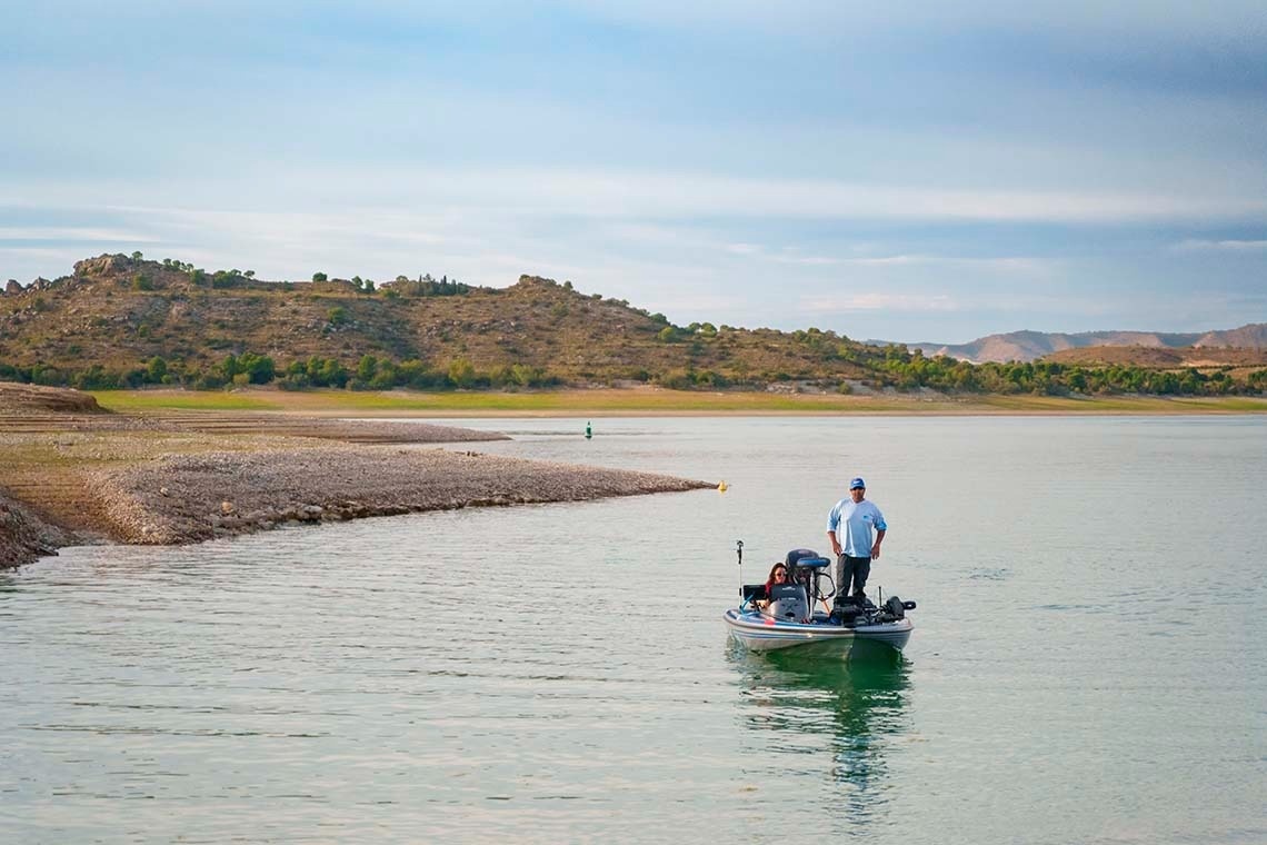Dos personas están en una lancha de pesca en un embalse con orillas rocosas expuestas debido al bajo nivel del agua y colinas áridas al fondo, bajo un cielo parcialmente nublado.