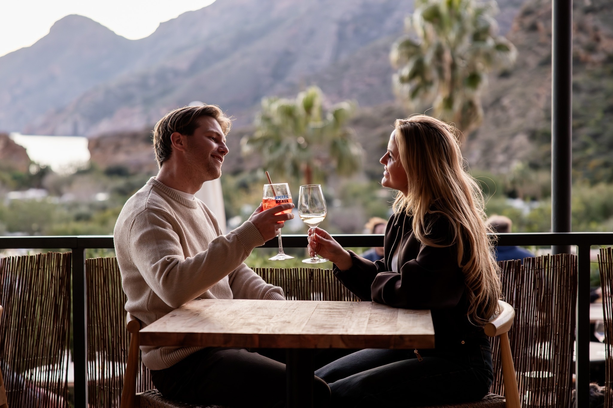 Una pareja sonriente brinda con copas en una terraza con montañas y el mar de fondo.