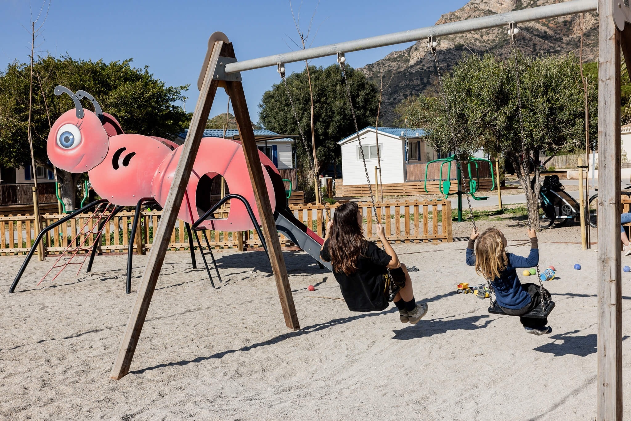 Dos niños se columpian en un parque de arena con una estructura de juego de hormiga roja, casas y montañas al fondo.