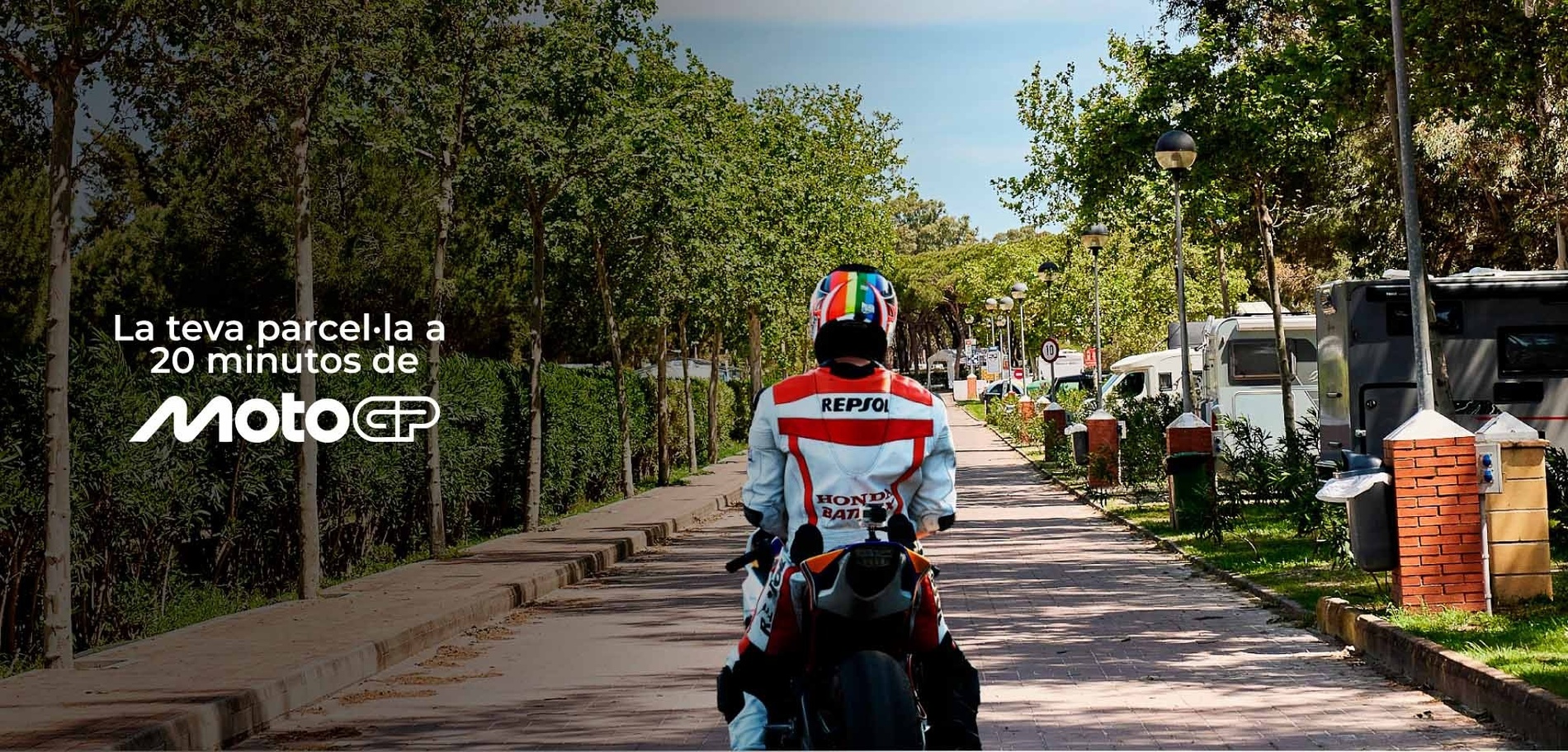 Un motorista amb vestit de Repsol i casc vibrant transita un camí arbrat amb caravanes, mentre un text promociona "La teva parcel·la a 20 minutos de MotoGP".