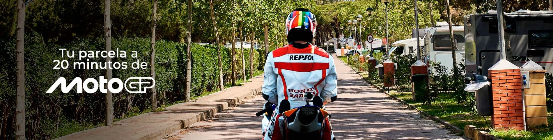 Un motociclista con traje de Repsol se aleja por un sendero arbolado en un camping, con el mensaje promocional "Tu parcela a 20 minutos de MotoGP".