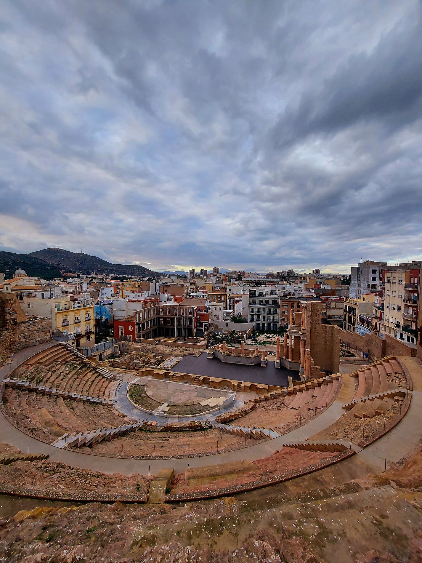 Taiga: Corporativa: Teatro Romano, Cartagena. Vista panorámica de las ruinas antiguas y la ciudad moderna bajo un cielo nublado.