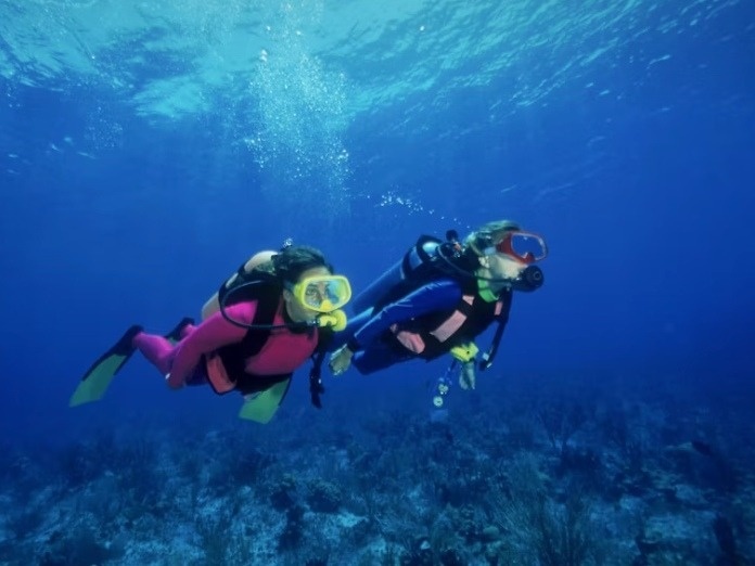Dos buceadores exploran el azulado fondo marino, con burbujas ascendiendo hacia la superficie.