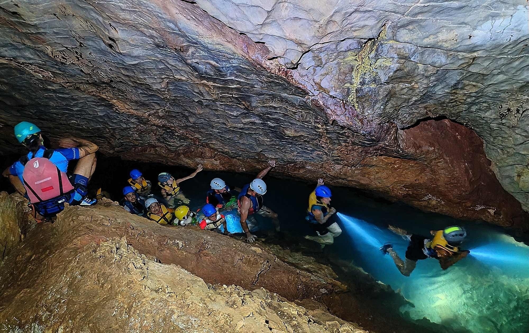 Varias personas disfrutan del kayak y la natación en aguas turquesas y claras junto a imponentes acantilados rocosos.