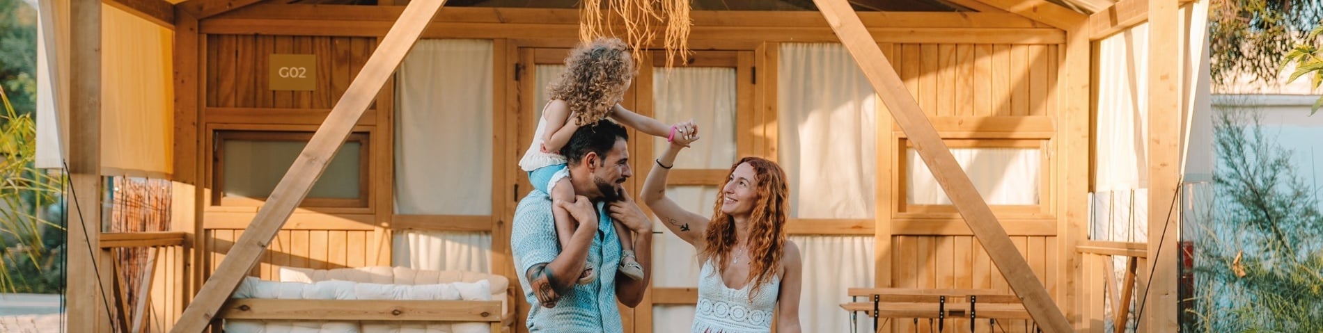 Een gelukkig gezin met een kind op de schouders lacht voor een houten accommodatie met de aanduiding 'G02'.