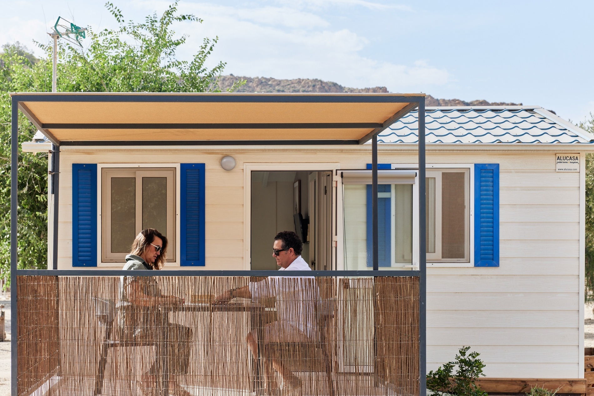 a man and a woman are sitting on a balcony in front of a white house with blue shutters