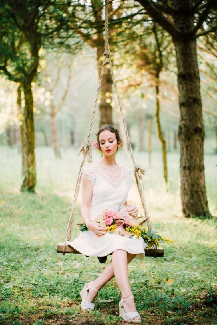 a woman in a white dress is sitting on a swing holding flowers