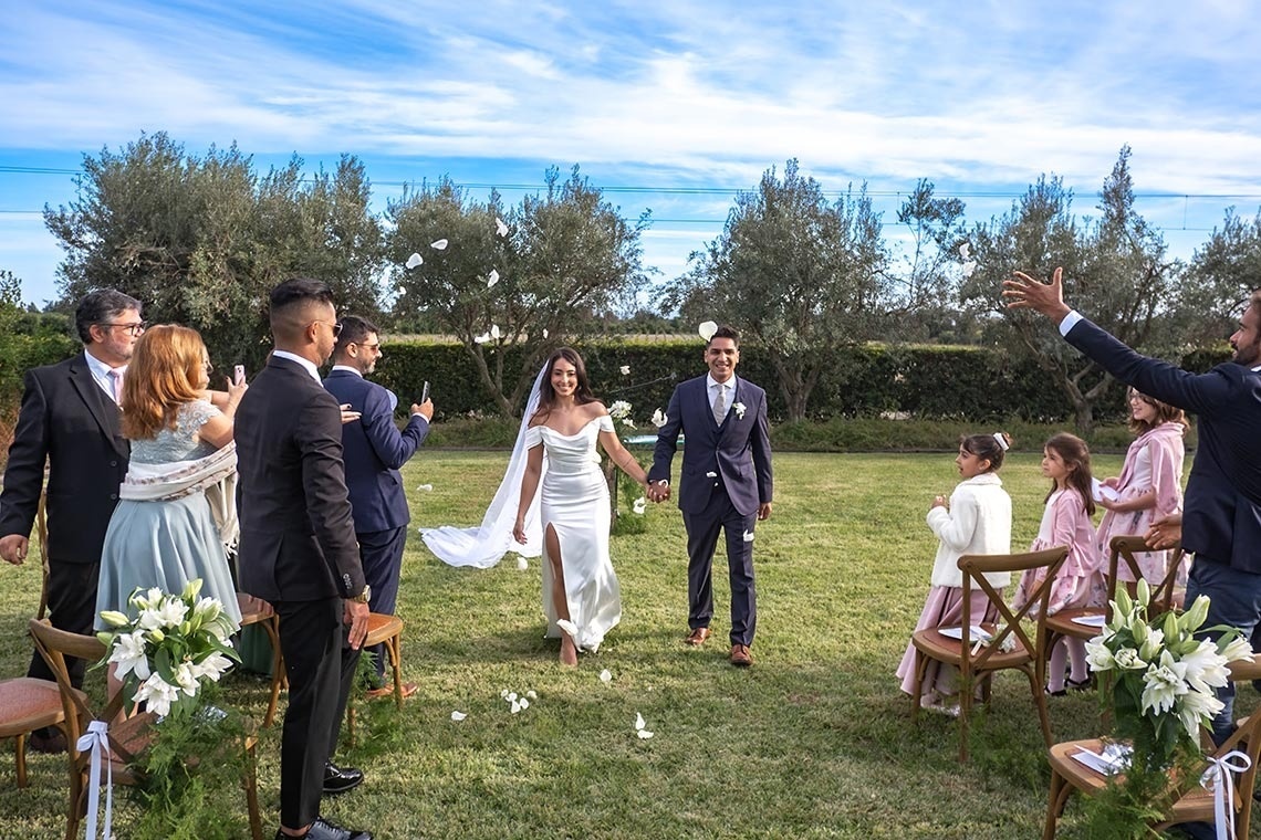 Um casal de noivos sorridente caminha de mãos dadas por um gramado, enquanto convidados jogam pétalas brancas em uma celebração ao ar livre.