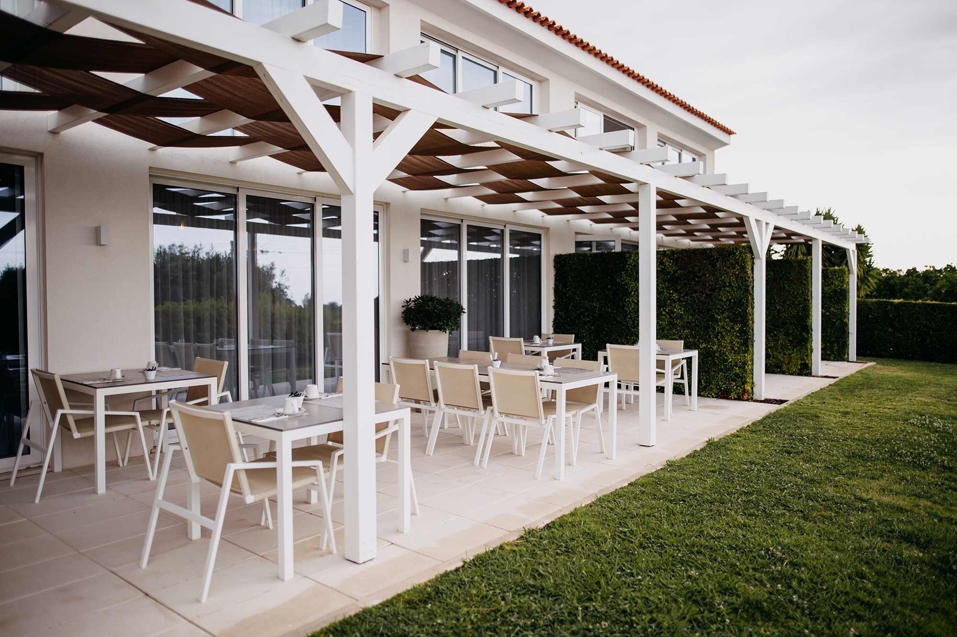 Une terrasse meublée de tables et de chaises, abritée par une pergola blanche et marron, longe un bâtiment clair et une pelouse verdoyante avec des haies.