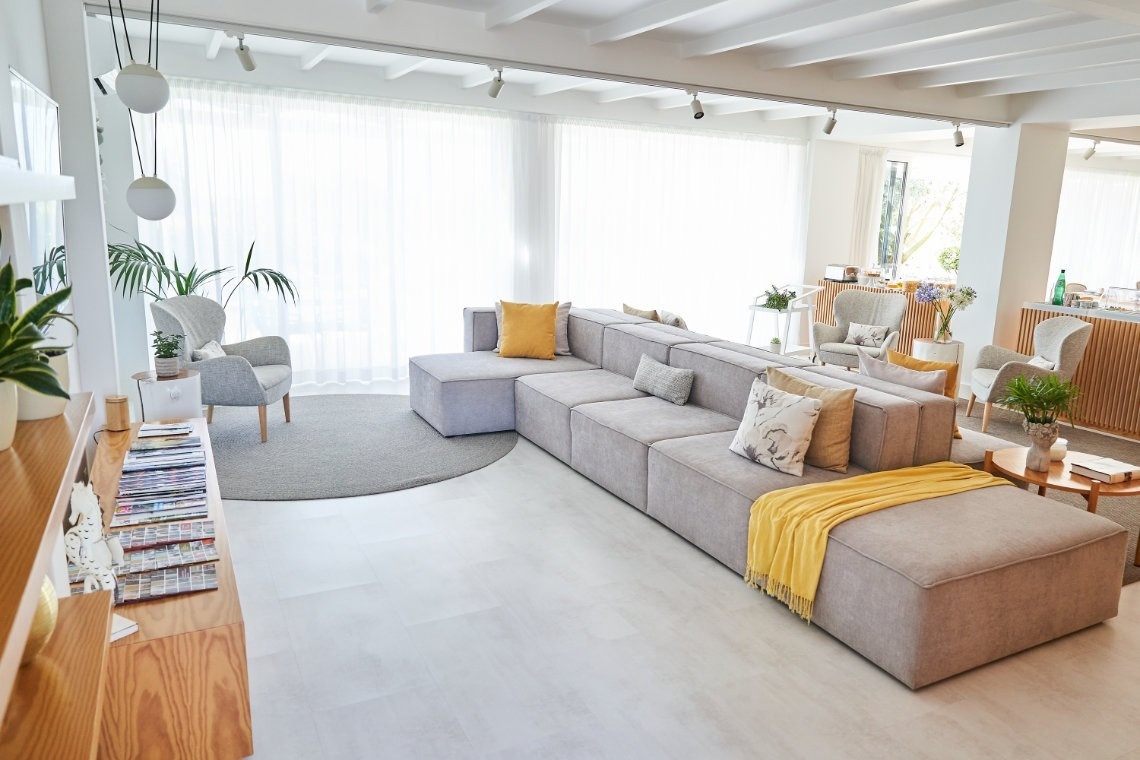 Un amplio y moderno salón presenta un gran sofá modular gris en forma de L con acentos amarillos, grandes ventanales con cortinas translúcidas y una decoración luminosa con plantas.