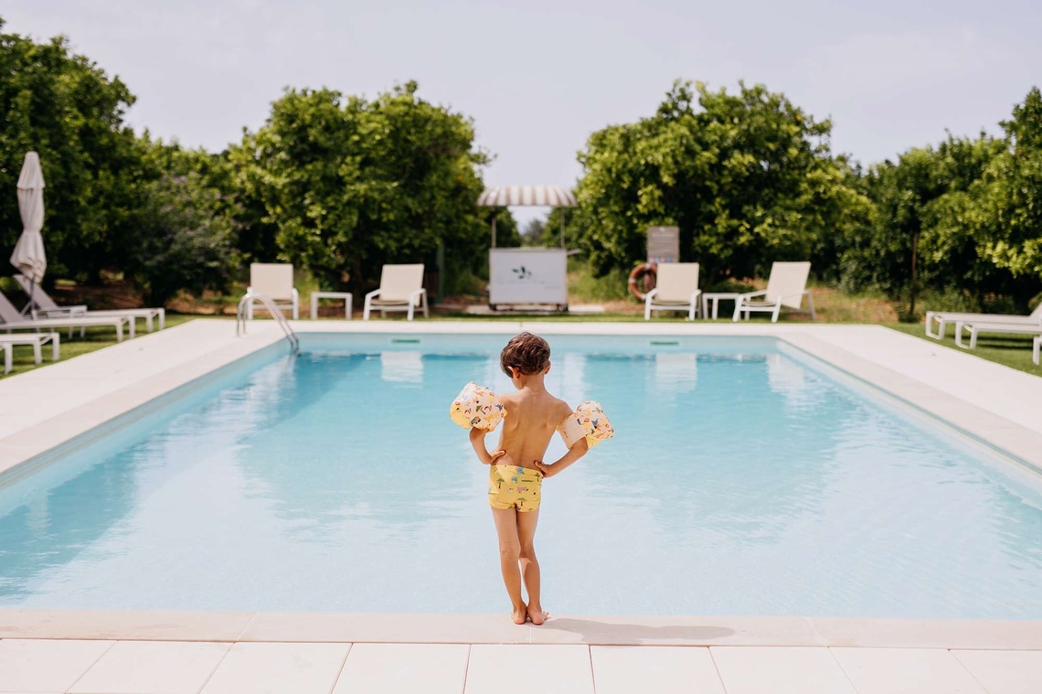 A young child wearing arm floats and swim trunks stands at the edge of a bright blue swimming pool, looking towards a lush green background with lounge chairs and a small snack cart.