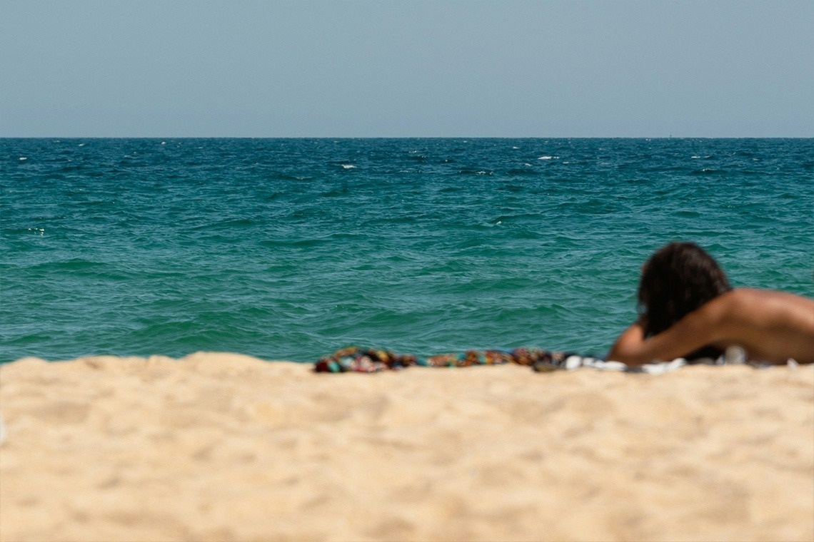Três pequenas cabanas amarelas se destacam à beira do mar azul, com uma cerca de madeira e vegetação verde e amarela em primeiro plano, sob um céu claro.