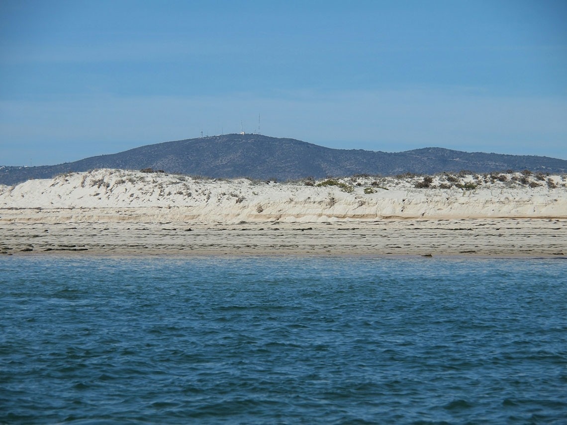 A imagem mostra uma paisagem costeira com águas azuis em primeiro plano, dunas de areia claras com vegetação e montanhas com torres ao fundo sob um céu azul.