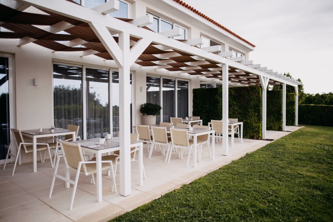 Una terraza de comedor exterior con mesas y sillas bajo una pérgola blanca, adyacente a un edificio y un jardín con césped y setos.