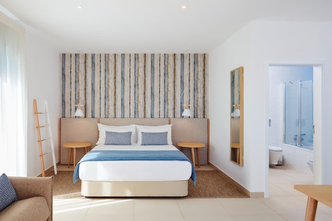 Un luminoso y moderno dormitorio de hotel con una cama doble, un cabecero decorado con franjas verticales de madera clara y azul, y un baño visible a través de una puerta abierta.