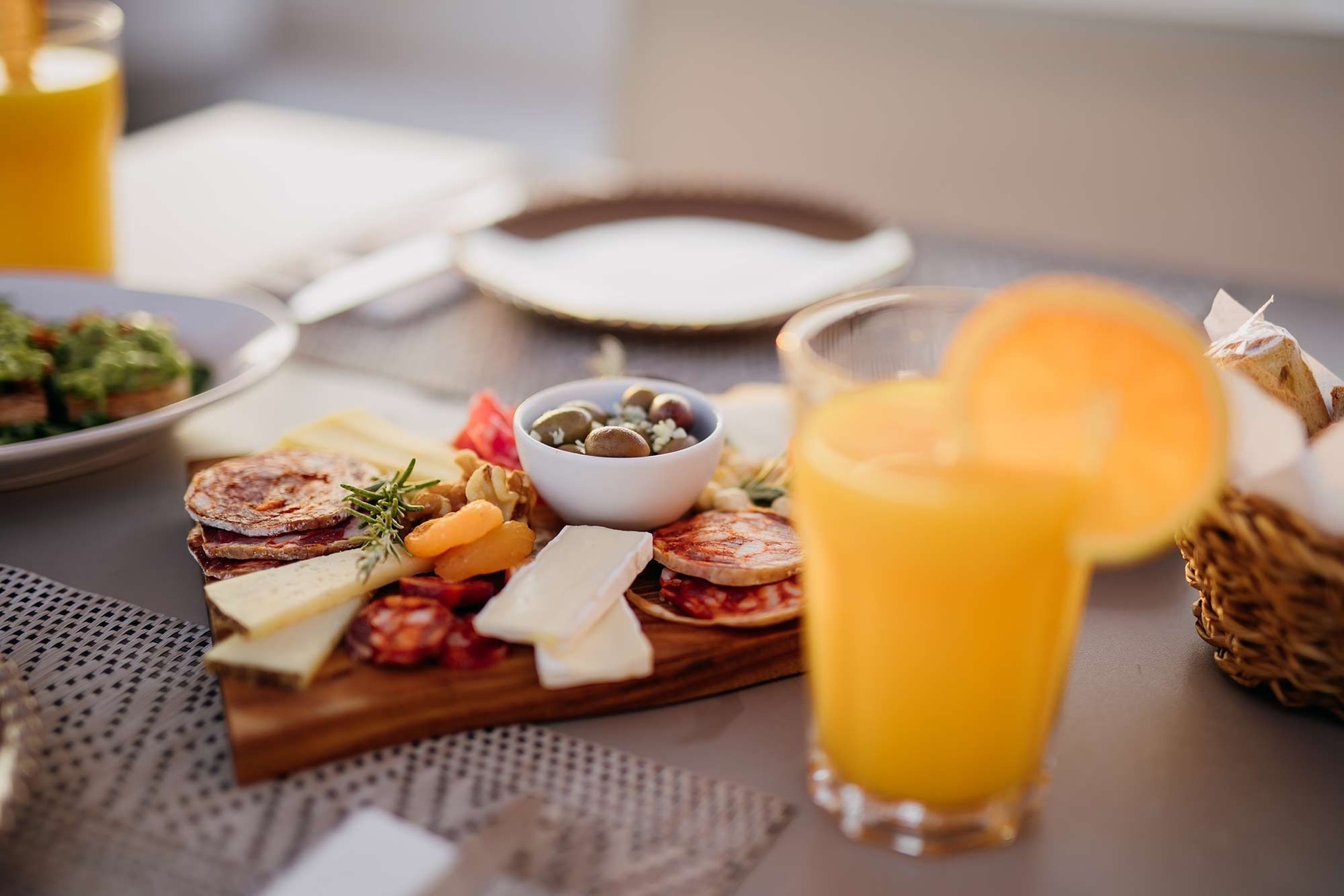 A wooden board laden with various cheeses, cured meats, nuts, and olives is prominently displayed on a table, with a glass of orange juice garnished with an orange slice in the foreground and other blurred dishes in the background.