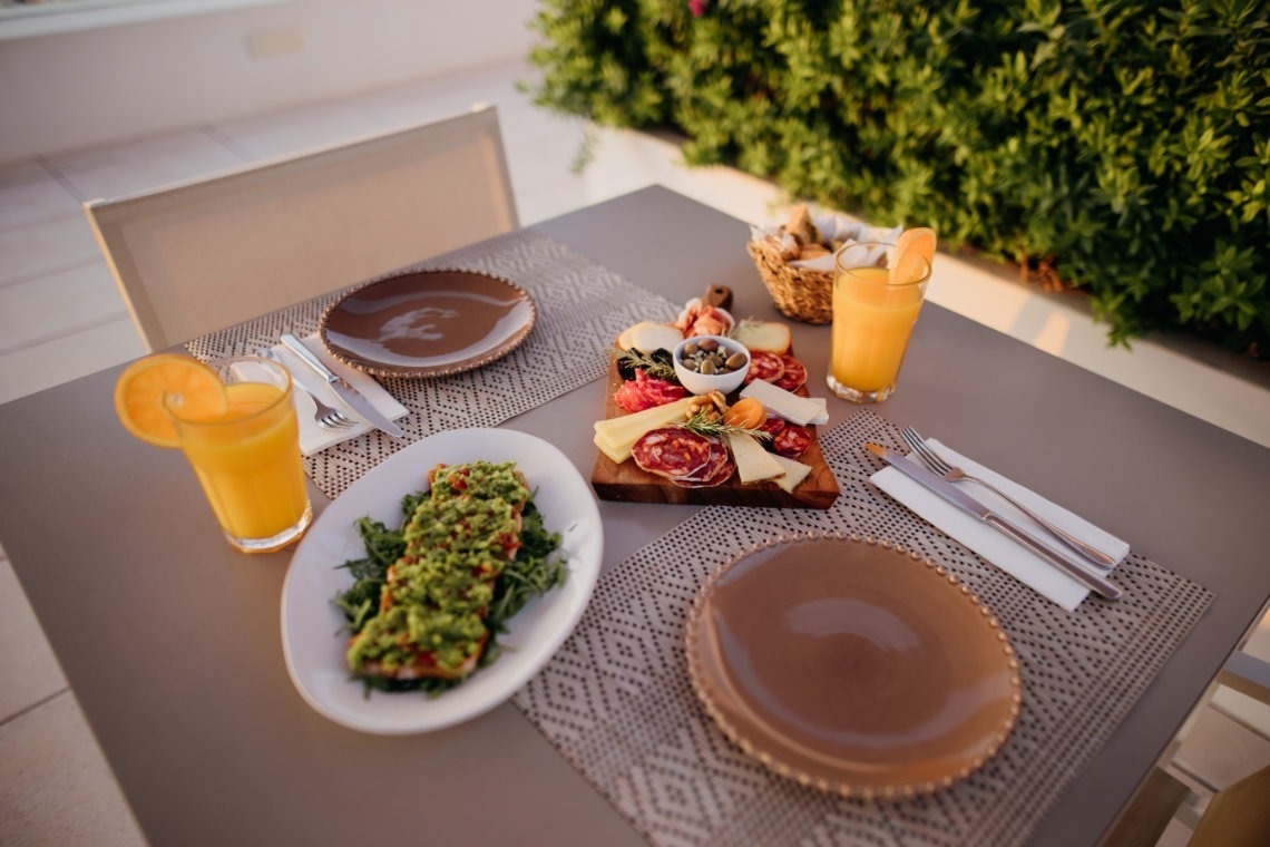 Uma mesa elegantemente posta ao ar livre apresenta uma tábua de frios, torradas com abacate e sumos de laranja, pronta para uma refeição para duas pessoas.