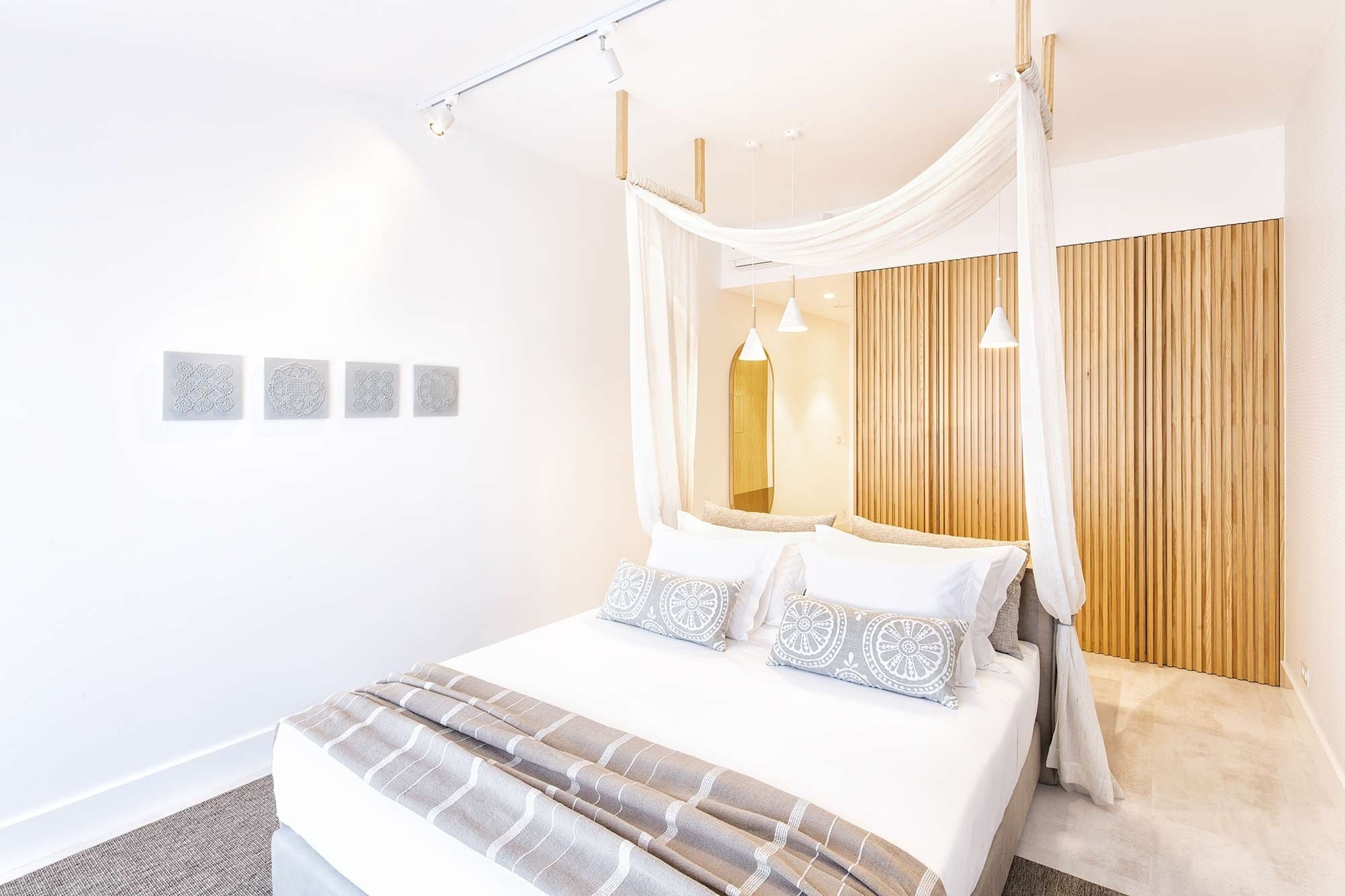 A bright, minimalist bedroom features a bed draped with a light fabric canopy, white bedding, decorative grey pillows, and a prominent wall covered in vertical wooden slats.
