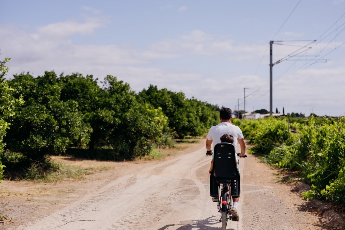 Um adulto pedala uma bicicleta com uma criança numa cadeira traseira, numa estrada de terra rodeada por vegetação e árvores, com postes de energia ao fundo.